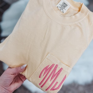 May include: A light yellow t-shirt with a pocket featuring pink embroidered initials. The shirt is folded, with the pocket detail visible. The tag reads "Comfort Colors".