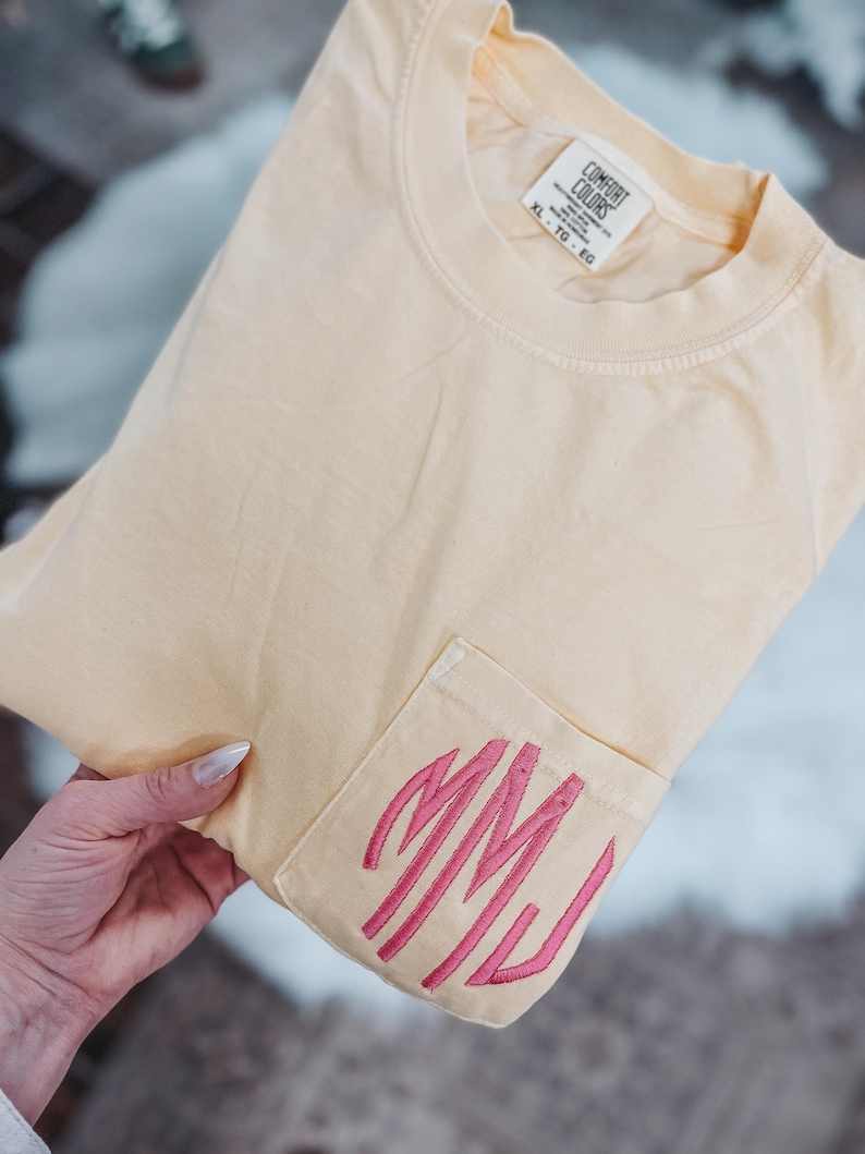 May include: A light yellow t-shirt with a pocket featuring pink embroidered initials. The shirt is folded, with the pocket detail visible. The tag reads "Comfort Colors".
