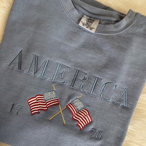 May include: A light blue sweatshirt with the word "AMERICA" embroidered in a matching blue thread. Below the word are two embroidered American flags with the numbers "17" and "76" on either side.
