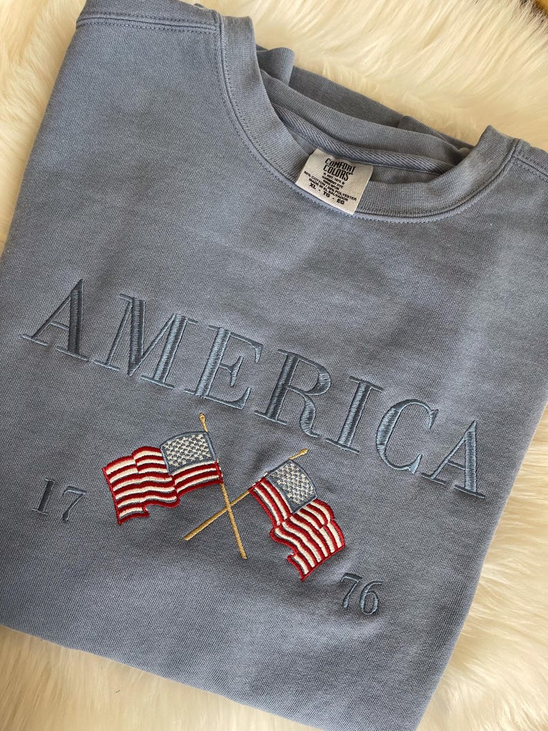 May include: A light blue sweatshirt with the word "AMERICA" embroidered in a matching blue thread. Below the word are two embroidered American flags with the numbers "17" and "76" on either side.