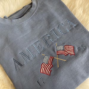 May include: A light blue sweatshirt with the embroidered word "AMERICA" and two crossed American flags. The numbers "17" and "76" are embroidered below and to the right of the flags. The sweatshirt is made of a soft material.