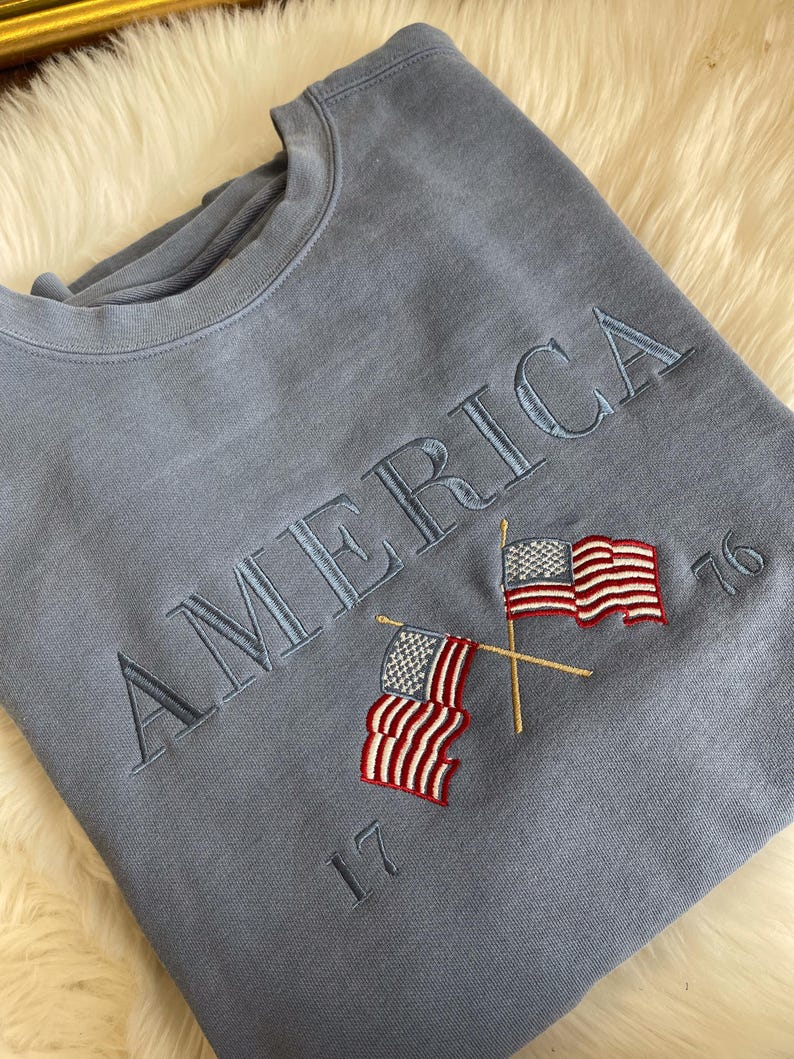 May include: A light blue sweatshirt with the embroidered word "AMERICA" and two crossed American flags. The numbers "17" and "76" are embroidered below and to the right of the flags. The sweatshirt is made of a soft material.