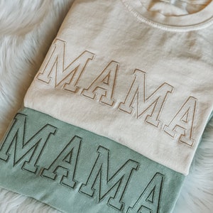 May include: Two t-shirts stacked on top of each other. The top shirt is white with the word "MAMA" embroidered in a light brown colour. The bottom shirt is a light green colour with the word "MAMA" embroidered in a dark green colour.