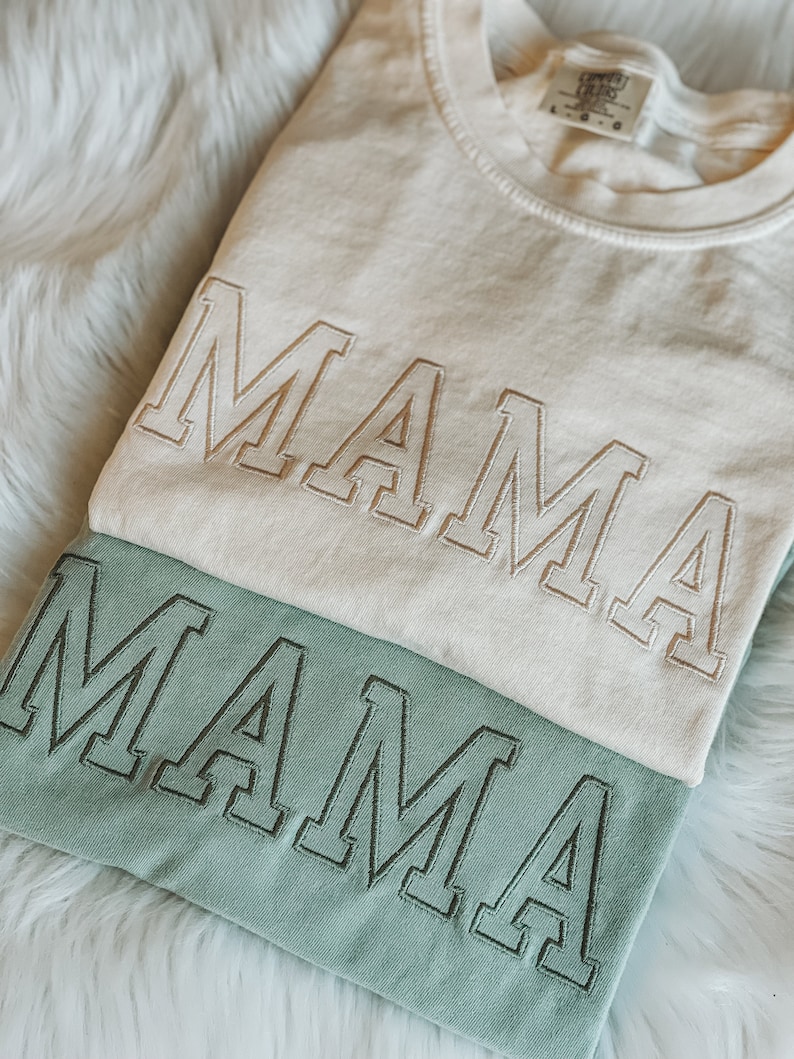 May include: Two t-shirts stacked on top of each other. The top shirt is white with the word "MAMA" embroidered in a light brown colour. The bottom shirt is a light green colour with the word "MAMA" embroidered in a dark green colour.