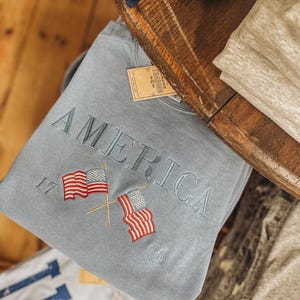 May include: A light blue sweatshirt with the word "AMERICA" embroidered in gray. Two crossed American flags are embroidered below the word, with the numbers "17" and "76" on either side. The sweatshirt is folded on a wooden surface.