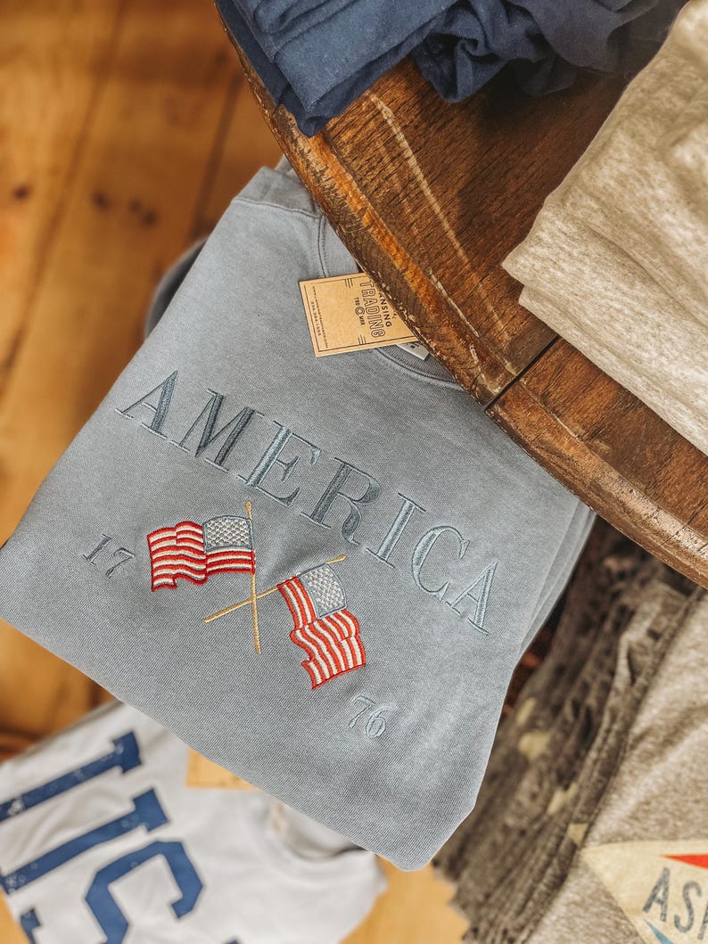 May include: A light blue sweatshirt with the word "AMERICA" embroidered in gray. Two crossed American flags are embroidered below the word, with the numbers "17" and "76" on either side. The sweatshirt is folded on a wooden surface.
