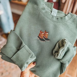 May include: A sage green crewneck sweatshirt featuring an embroidered design of two brown and black duck boots on the center chest. The garment has long sleeves with ribbed cuffs and a soft fleece interior lining visible at the folded sleeve edges.