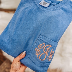 May include: A blue Comfort Colors t-shirt with a pocket featuring an embroidered orange monogram. The shirt is folded, showing the pocket detail. The shirt has a crew neck and a tag visible at the neckline.