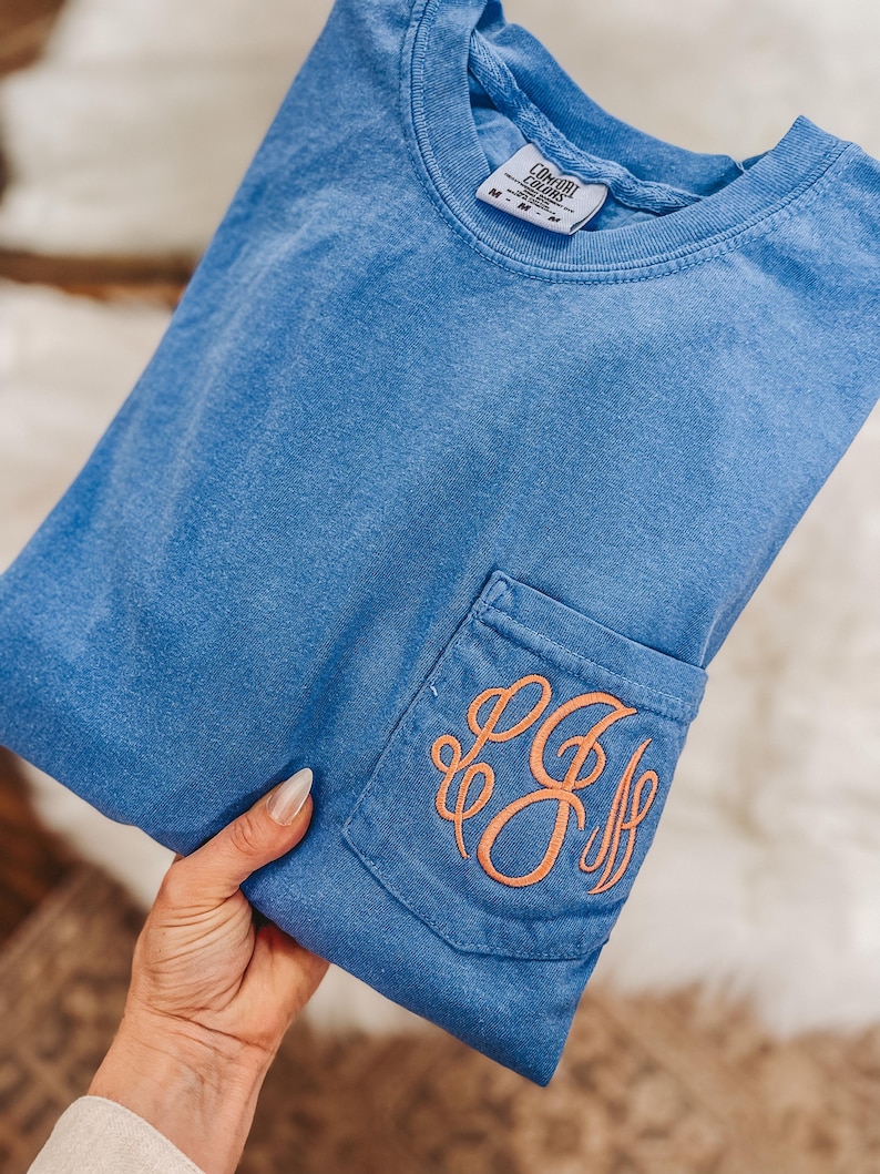 May include: A blue Comfort Colors t-shirt with a pocket featuring an embroidered orange monogram. The shirt is folded, showing the pocket detail. The shirt has a crew neck and a tag visible at the neckline.