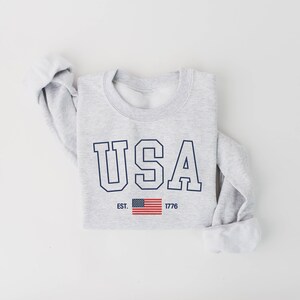 May include: A gray sweatshirt with the word "USA" printed in navy blue letters with a white outline. The letters are in a bold, blocky font. Below the word "USA" is a small American flag and the text "EST. 1776".