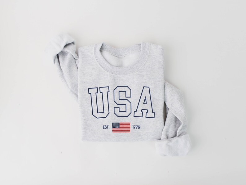 May include: A gray sweatshirt with the word "USA" printed in navy blue letters with a white outline. The letters are in a bold, blocky font. Below the word "USA" is a small American flag and the text "EST. 1776".