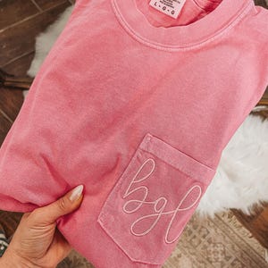 May include: A pink short-sleeved t-shirt with a crew neckline and a single chest pocket. The pocket features embroidered cursive letters that read bgl. The interior neck label displays the text Comfort Colors, Heavyweight Garment Dye, Ring Spun, 100% Cotton, Made in Honduras, and size L-G-G.