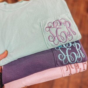 May include: A stack of three t-shirts in pastel colors: mint green, purple, and pink. Each shirt has a pocket with embroidered initials in coordinating colors. The top shirt has purple embroidery, the middle shirt has teal, and the bottom shirt has a light brown.
