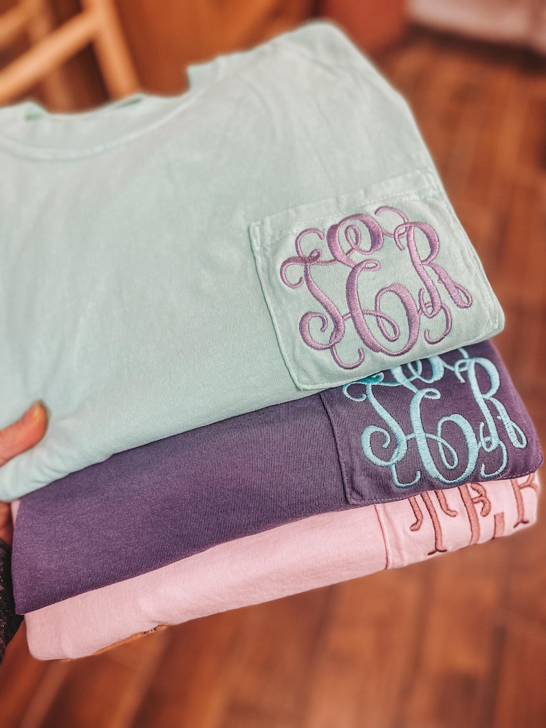 May include: A stack of three t-shirts in pastel colors: mint green, purple, and pink. Each shirt has a pocket with embroidered initials in coordinating colors. The top shirt has purple embroidery, the middle shirt has teal, and the bottom shirt has a light brown.