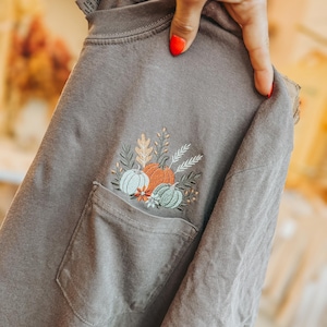 May include: A charcoal gray t-shirt featuring a chest pocket with an embroidered design of three pumpkins in shades of orange, light blue, and sage green, accented with wheat stalks, small flowers, and autumn leaves arranged above the pocket.