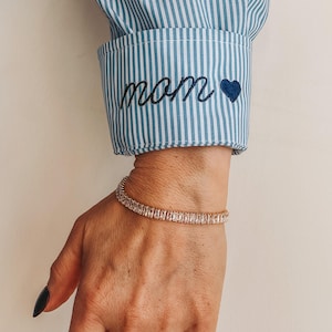 May include: A gold-toned bracelet with rectangular clear stones is worn on a wrist. The cuff of a blue and white striped shirt is visible, embroidered with the word "mom" and a blue heart.