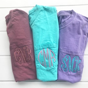 May include: Three folded t-shirts in burgundy, turquoise, and lavender, each with a pocket embroidered with a monogram. The shirts are displayed on a white wooden surface.