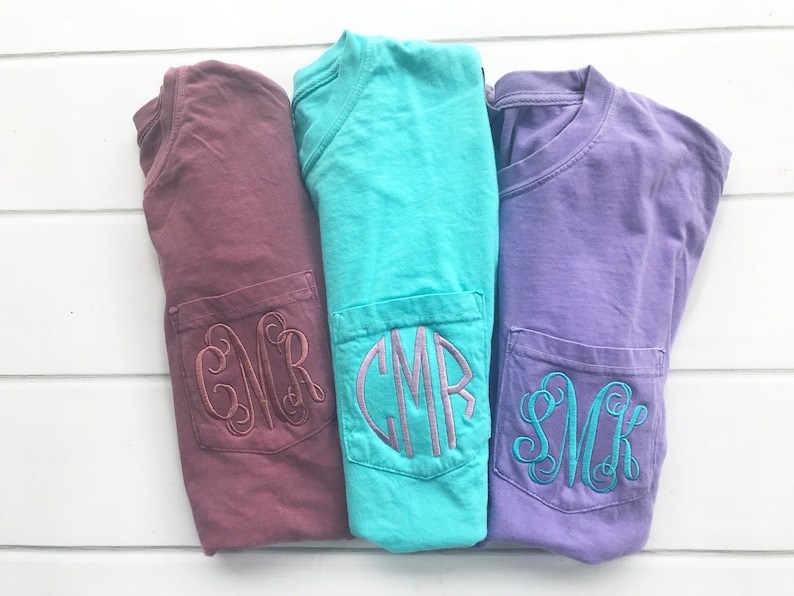 May include: Three folded t-shirts in burgundy, turquoise, and lavender, each with a pocket embroidered with a monogram. The shirts are displayed on a white wooden surface.