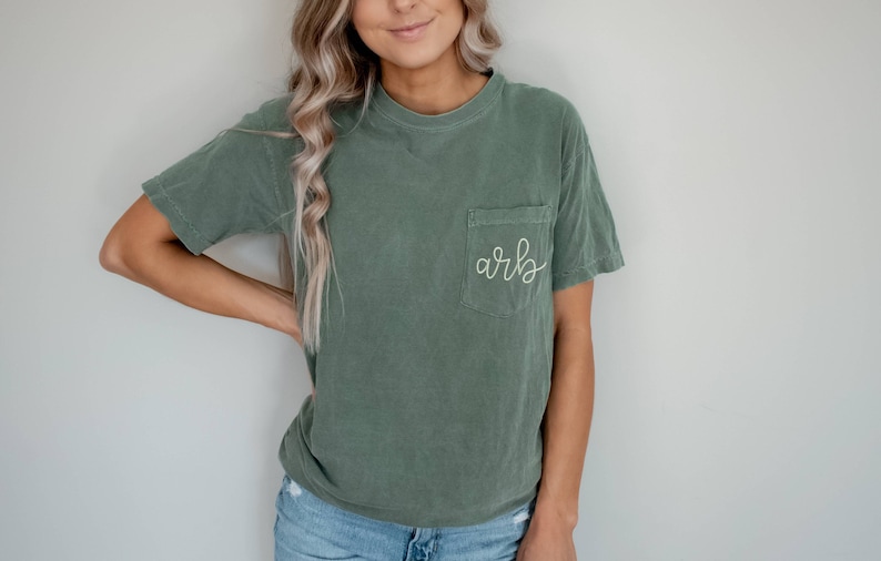May include: Olive green short-sleeved t-shirt with a pocket on the chest. The pocket features the embroidered initials "arb" in a cursive font. The shirt is made of a soft, comfortable material.