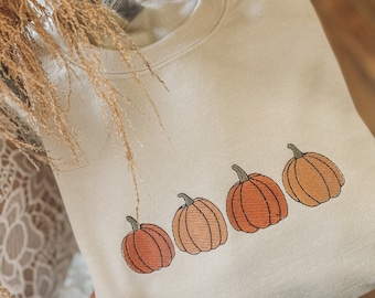 Pode incluir: Uma camisa de moletom bege com uma linha de quatro abóboras bordadas em tons de laranja e marrom.