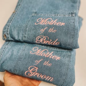 Op de afbeelding: Twee lichtblauwe spijkeroverhemden met roze geborduurde tekst. Eén overhemd leest "Mother of the Bride" en de andere "Mother of the Groom". De overhemden zijn opgevouwen en lijken nieuw.