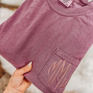 May include: A mauve-colored pocket t-shirt with the embroidered initials "MM" on the pocket. The shirt is a Comfort Colors brand, size XL, and has a crew neck. The fabric appears soft and slightly faded.