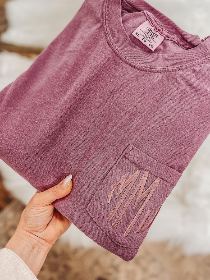 May include: A mauve-colored pocket t-shirt with the embroidered initials "MM" on the pocket. The shirt is a Comfort Colors brand, size XL, and has a crew neck. The fabric appears soft and slightly faded.