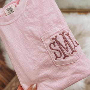 May include: A light pink Comfort Colors t-shirt with a pocket featuring embroidered initials. The shirt is folded, showcasing the pocket detail with the initials in a darker shade of pink. The Comfort Colors tag is visible at the neckline.