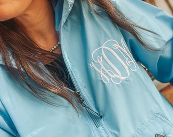May include: A pale blue anorak jacket with a quarter-zip and a front pocket. The jacket has a white embroidered monogram on the chest. The anorak features a hood and a zipper pull. The jacket is made of a lightweight material.
