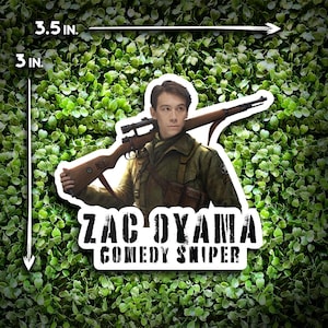 May include: A sticker featuring a man in a military uniform holding a rifle. The sticker is 8.89 cm wide and 7.62 cm tall. The text "ZAC OYAMA COMEDY SNIPER" is printed in bold, black letters below the image.