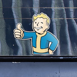 Puede incluir: Una pegatina con el personaje Vault Boy de la serie de videojuegos Fallout. El personaje se representa con pelo rubio, un mono azul y un gesto de pulgar hacia arriba. La pegatina tiene un borde blanco.