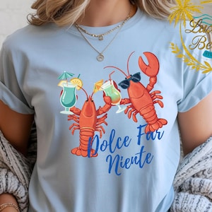 May include: A light blue t-shirt featuring a graphic of two orange lobsters holding tropical cocktails with umbrellas. One lobster wears black sunglasses. The text "Dolce Far Niente" is printed in blue script below the lobsters. The shirt has short sleeves.