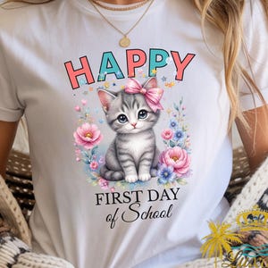Puede incluir: Una camiseta blanca con un gráfico de un gatito atigrado gris con un lazo rosa, rodeado de flores y estrellas de colores. El texto dice "HAPPY" en letras color coral sobre el gatito y "FIRST DAY of School" en letra cursiva negra debajo.