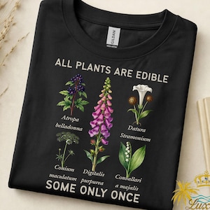 May include: A black t-shirt featuring the white text ALL PLANTS ARE EDIBLE SOME ONLY ONCE. The graphic includes botanical illustrations of Atropa belladonna, Conium maculatum, Digitalis purpurea, Datura stramonium, and Convallaria majalis with labels.