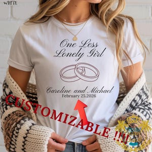 Peut inclure: T-shirt blanc avec l'inscription "One Less Lonely Girl" au-dessus de deux anneaux entrelacés. Les noms "Caroline and Michael" et la date "February 25, 2026" sont imprimés en dessous. Le mot "CUSTOMIZABLE" est imprimé en rouge.