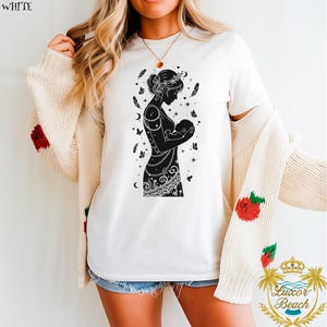 以下が含まれることがあります： 白いTシャツ。黒いシルエットで、母親が赤ちゃんに母乳を与えている様子が描かれています。デザインには羽、蝶、三日月などの装飾要素が含まれています。Tシャツは半袖で、クリーム色のカーディガンと合わせて着用されています。