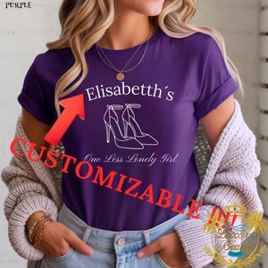 Peut inclure: T-shirt violet avec le texte "Elisabetth's" au-dessus d'un dessin de deux chaussures à talons hauts. En dessous, le texte "One Less Lonely Girl". Le mot "CUSTOMIZABLE" est imprimé en rouge sur le devant du t-shirt.