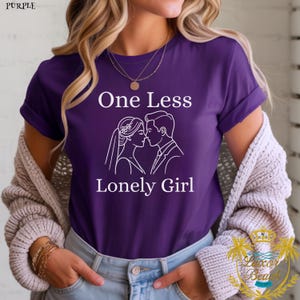 Peut inclure: T-shirt violet avec l'inscription "One Less Lonely Girl" et un dessin au trait blanc d'un couple s'embrassant. Le t-shirt a des manches retroussées et est porté.