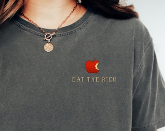 May include: Dark gray t-shirt with the phrase "EAT THE RICH" in gold lettering and a red apple graphic. The shirt is a crew neck and has short sleeves. The shirt is made of a soft, comfortable material.