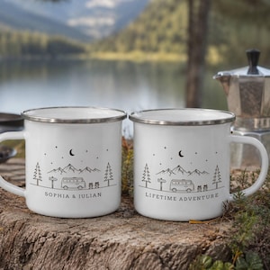 Könnte beinhalten: Zwei weiße Emaille-Tassen mit silbernem Rand, jeweils mit einer schwarzen Strichzeichnung eines Campingbusses, Bergen, Bäumen und einer Mondsichel. Eine Tasse ist mit "SOPHIA & JULIAN" und die andere mit "LIFETIME ADVENTURE" beschriftet.