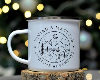 May include: A white enamel mug with a silver rim featuring a black circular graphic of mountains, a tent, a winding path, and a crescent moon. The text reads VIVIAN & MATTIAS and LIFETIME ADVENTURE with EST. 2026 in the center of the design.