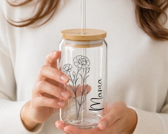 May include: A clear glass tumbler with a bamboo lid and a clear straw. The tumbler features a black floral design and the word "Mama" in script. The tumbler is held by a person wearing a white sweater.
