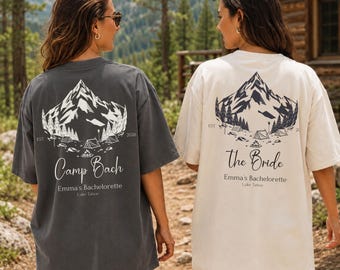 May include: Two short-sleeve t-shirts in pepper gray and ivory. The back of each shirt features a mountain and forest graphic with the text "EST 2026". The gray shirt reads "Camp Bach Emma's Bachelorette Lake Tahoe" and the ivory shirt reads "The Bride Emma's Bachelorette Lake Tahoe".