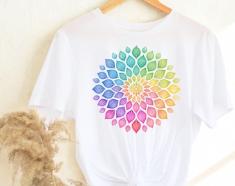 May include: White t-shirt featuring a colorful floral design. The design is a circular arrangement of leaf-shaped elements in a rainbow of colors, including red, orange, yellow, green, blue, and purple. The shirt is hanging on a wooden hanger.