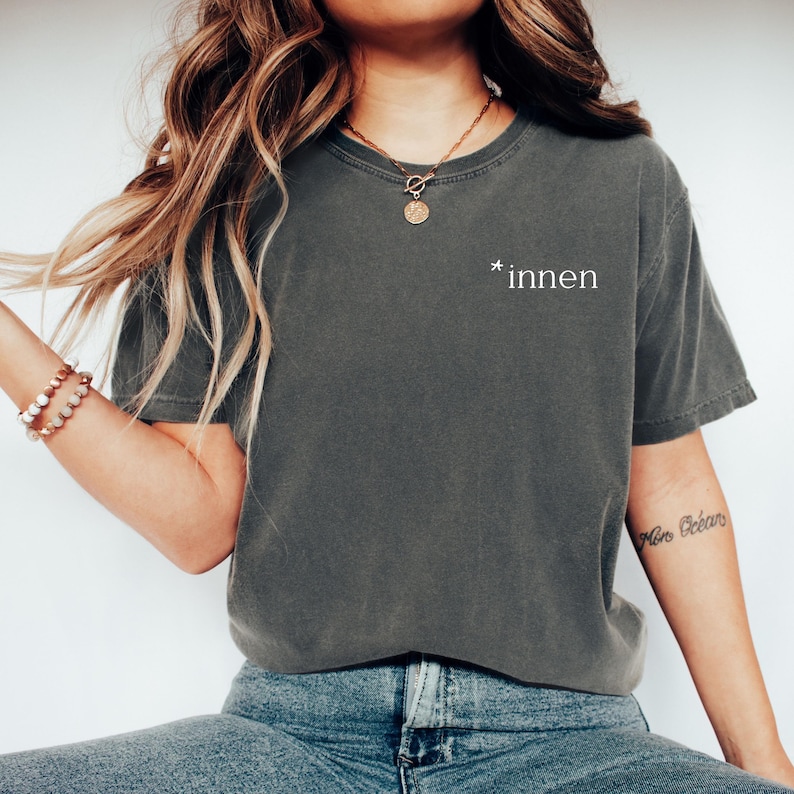 K&ouml;nnte beinhalten: Dunkelgraues T-Shirt mit dem wei&szlig;en Aufdruck "*innen" auf der Brust. Das Shirt hat kurze &Auml;rmel und einen Rundhalsausschnitt. Das Model tr&auml;gt eine blaue Jeans und eine goldene Halskette.