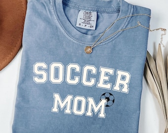 May include: A light blue t-shirt with the words "SOCCER MOM" in white block letters and a soccer ball graphic. The shirt has a crew neck and short sleeves. A gold necklace with a pendant is draped on the shirt.