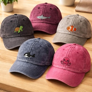 Könnte beinhalten: Fünf Baseballkappen in verschiedenen Farben, darunter Dunkelgrau, Kastanienbraun, Hellgrau, Marineblau und Pink. Jede Kappe zeigt ein kleines gesticktes Meerestier: eine grüne Schildkröte, einen grauen Hai, einen orangefarbenen Clownfisch, einen schwarzen Orca und einen pinken Oktopus.