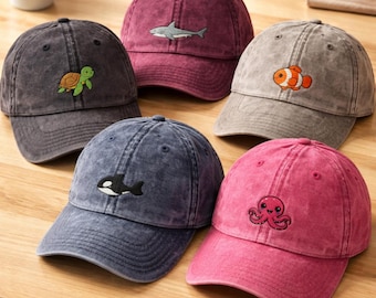 May include: Five baseball caps in various colors including dark gray, maroon, light gray, navy blue, and pink. Each cap features a small embroidered sea creature: a green turtle, a gray shark, an orange clownfish, a black orca, and a pink octopus.