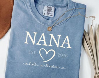 May include: A blue short-sleeved t-shirt featuring the white text NANA, EST. 2026, and a heart graphic. Below the heart, the names Nelli and Theodore are printed in a cursive script with small heart accents. A gold necklace rests on the shirt.