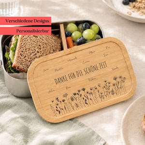 Könnte beinhalten: Eine rechteckige Edelstahl-Lunchbox mit einem Bambusdeckel. Der Deckel ist mit einem floralen Lasergravur-Muster, dem Text "DANKE FÜR DIE SCHÖNE ZEIT" und mehreren Namen versehen. Die Box enthält ein belegtes Brot sowie grüne Trauben und Blaubeeren.